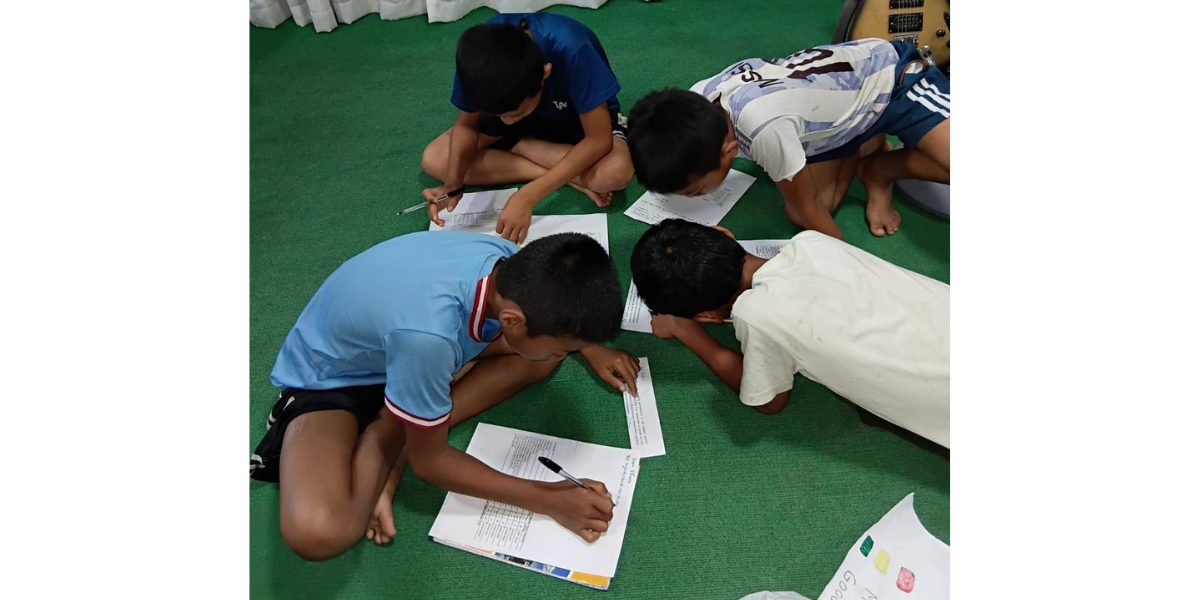 Four boys are sitting on the floor lined with green carpet, studying and writing together