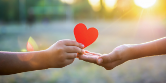 A hand passes a paper heart into an open hand, in front of a blurred background of a field with a setting sun.